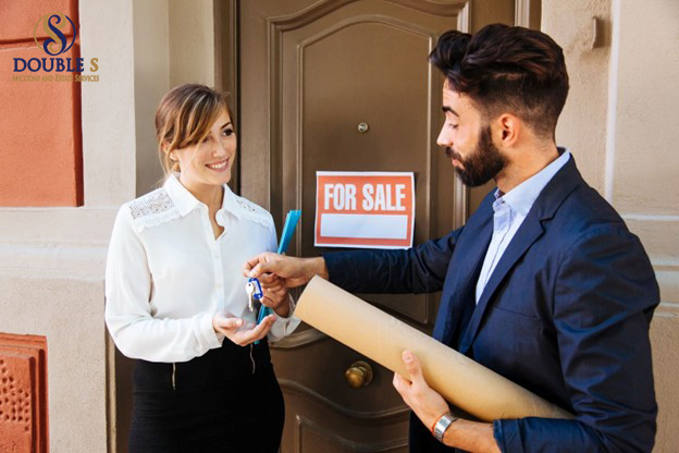 Agent giving house keys to a woman in front of a door with a for-sale sign.